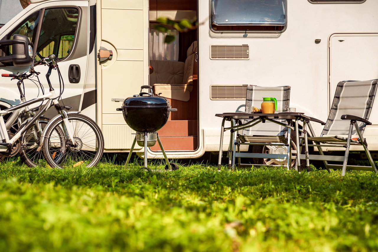 Vor einem Wohnmobil stehen Fahrräder, ein Grill und ein Campingtisch mit Campingstühlen.